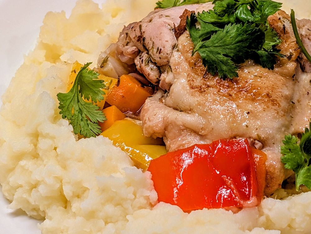 A close-up photo of mashed potatoes, vegetables and chicken topped with cilantro. The vegetables are carrots, red and yellow pepper. The chicken skin looks toasted. 