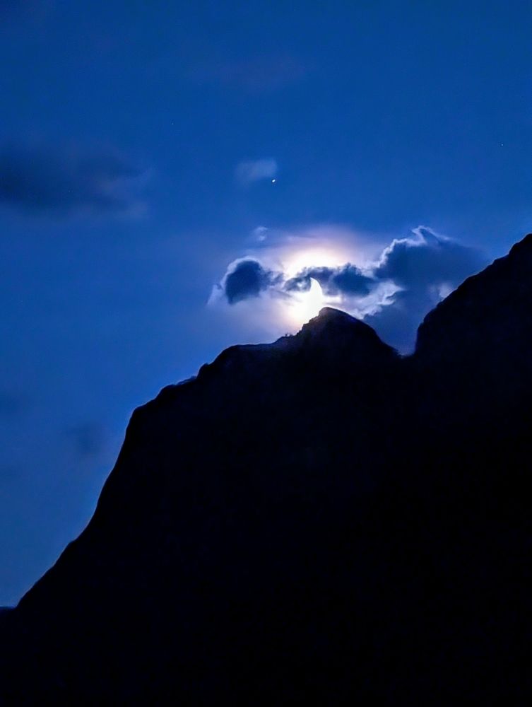 The moon is about to set behind the mountains in Glacier National Park in Montana. The black silhouette of the mountains is visible on the lower part of the photo, like a dark blue sky is seen on the top. Small clouds diffuse the light of the moon, and one star or planet is visible. 