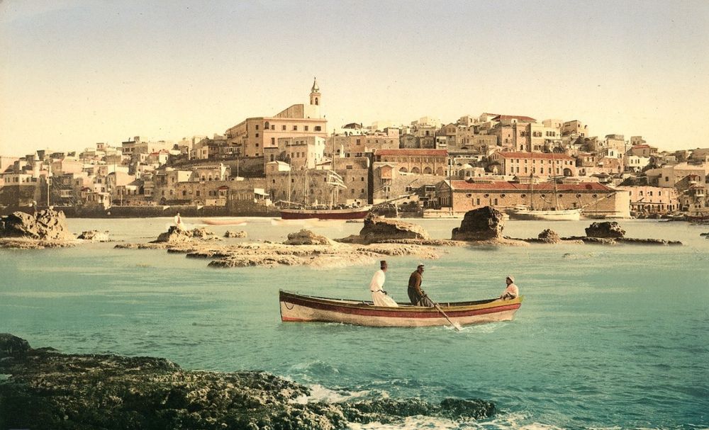 A view of a fishing boat in the harbour with the seafront of Jaffa in the background, including Mediterranean red tiled houses and a tower 