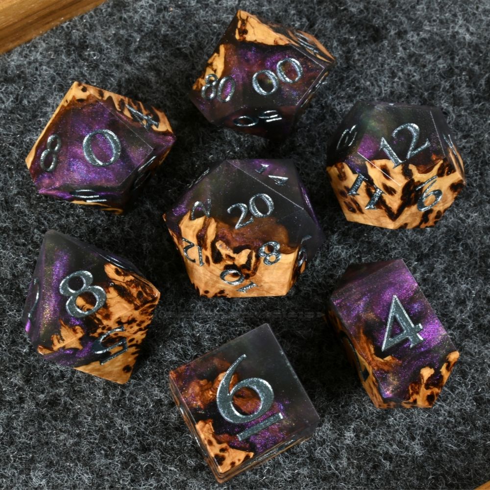 A set of polyhedral dice resting on  charcoal felt. The dice are made from York Gum burl wood and resin. The wood is various shades of redish brown with dark marbling. The resin is translucent black with swirling purple glitter that color shifts to blue, green, and gold. The numbers are painted dark silver.