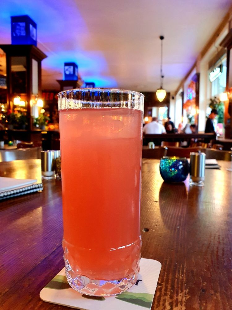 A tall glass of coral-pink sparkling cocktail sits on a wooden table in a warmly lit restaurant or bar. The textured crystal glass catches the ambient lighting, while blue decorative lights and other diners create a cozy atmosphere in the background. A small decorative sphere and napkin are visible on the table beside the drink.
