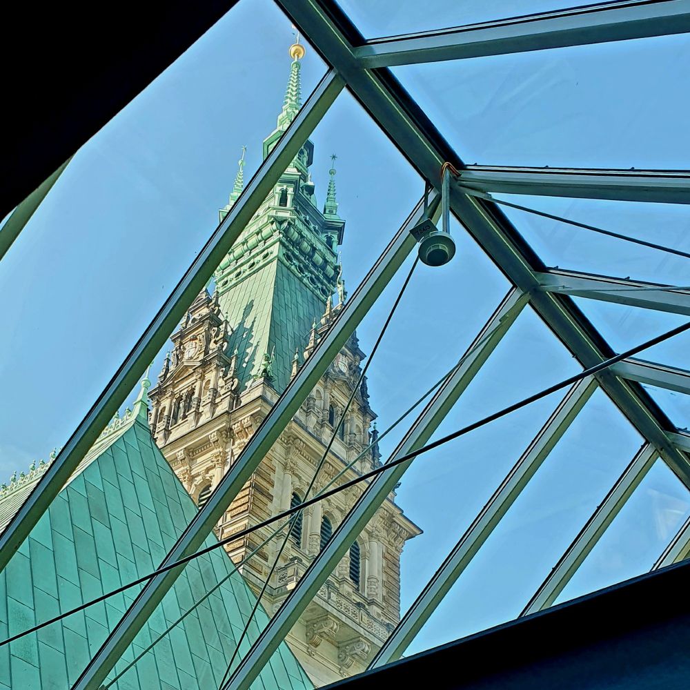 Looking up through a modern glass roof structure at the ornate tower of Hamburg's historic Rathaus (City Hall). The neo-Renaissance tower features copper-green roofing, decorative spires, and intricate stonework, framed dramatically by geometric glass panels and metal supports against a clear blue sky.
