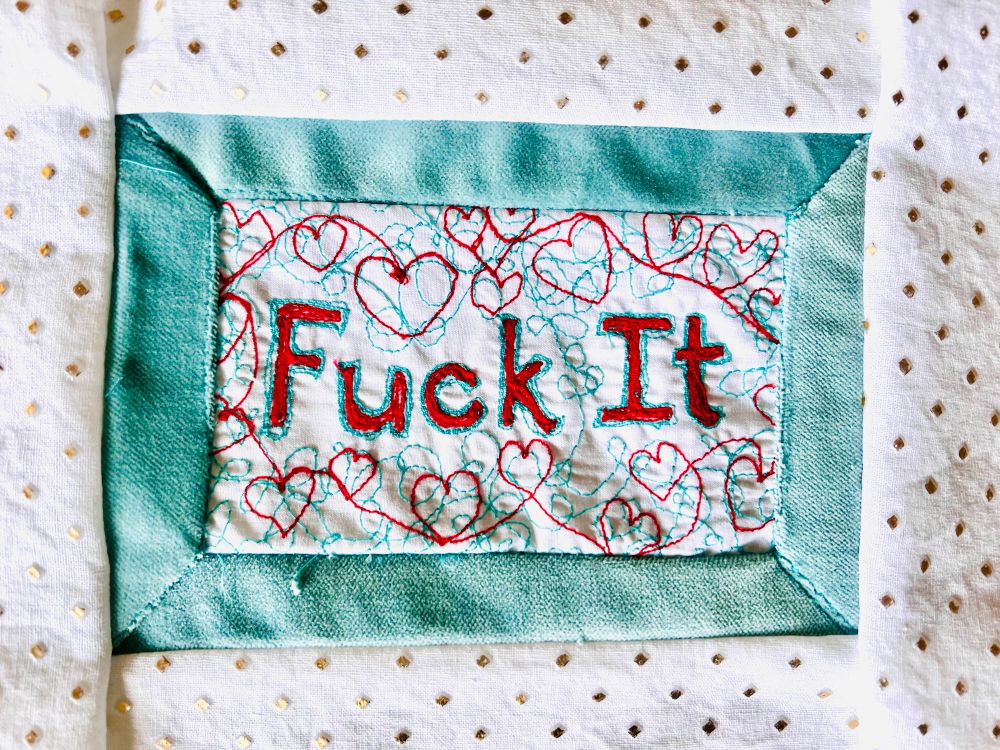 Red and teal “Fuck It” sewn onto white cloth and framed with teal velvet and a third white and gold fabric.