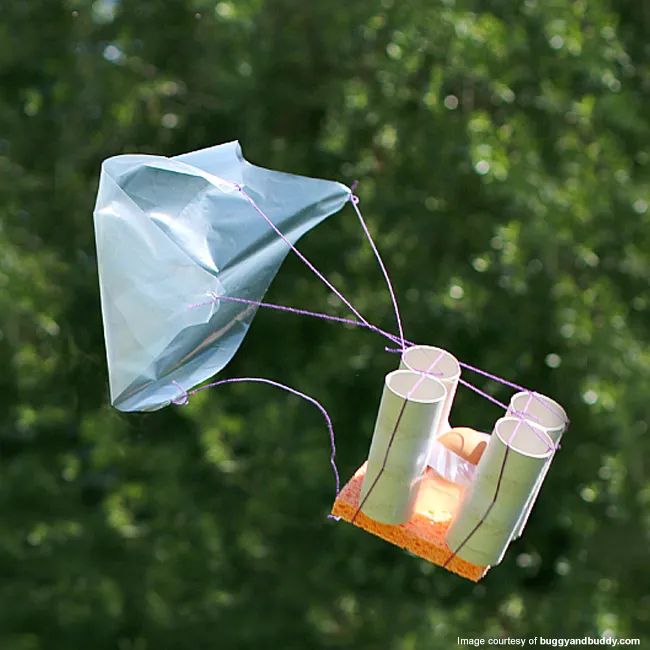 An egg cushioned by a kitchen sponge and toilet paper rolls, suspended from a paper parachute.