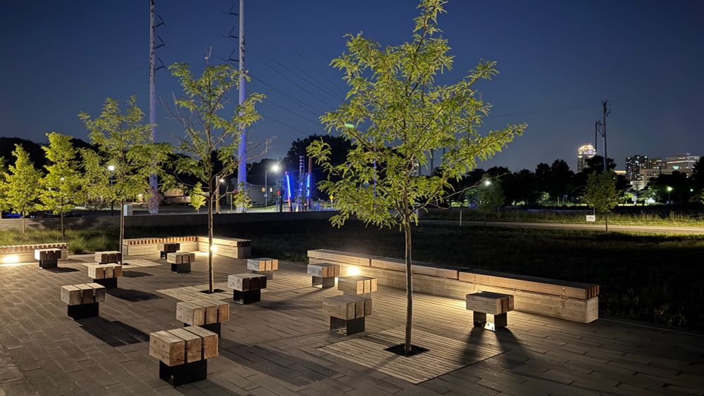 Graco park after dark in NE minneapolis. Lovely bench seating and landscape lighting. Newly planted young trees are showing green leaves and a clear night time sky in dark blue with some buildings visible on the horizon. This is right next to Mississippi River 