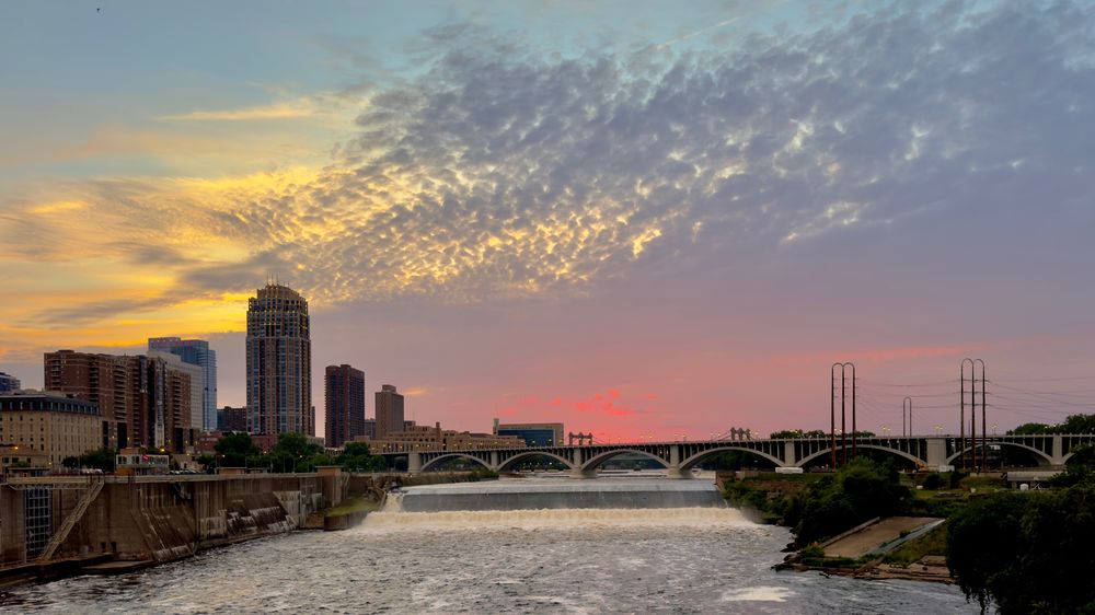 St Anthony Falls after dusk and the Mississippi River with a rainbow of colors visible in the clouds and sky. The view includes some skyline of minneapolis with lights amd the third avenue bridge showing clearly. 