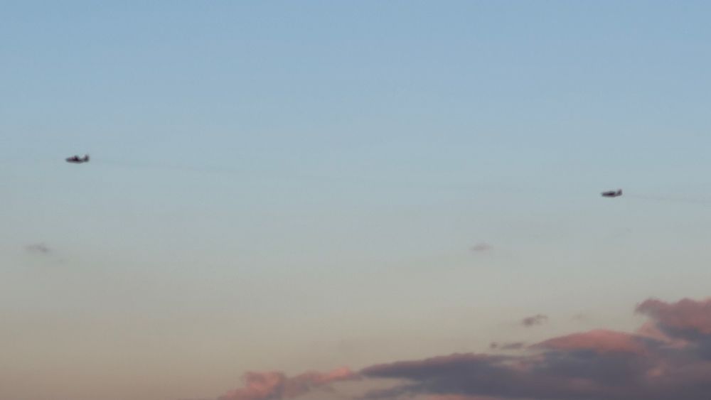 Two C130 in close formation after takeoff flying over NE minneapolis at less than 3.000 feet elevation. Sunset gradient colors from baby aqua blue to orangish neutral gray in a mostly clear sky with some dramatic clouds bottom right of the frame. 