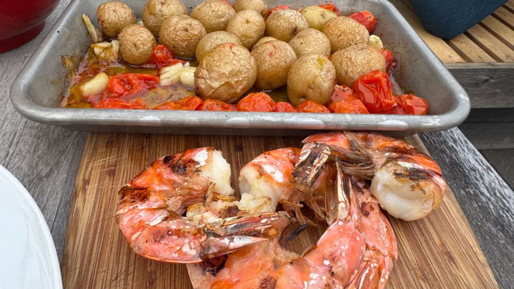 Cutting board with three large grilled shrimp and an eighth sheet pan full of with blistered cherry tomatoes, lovely small potatoes in olive oil with garlic and salt. 