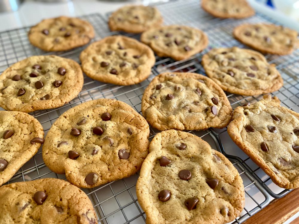 A wire rack cooling chocolate chip cookies on a white countertop.  The cookies are brown and slightly crispy on the edges and gooey & soft in the middle. 