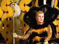 Alice Ghostley as Queen Bee is sitting - she is wearing a gold sequined long sleeve body suit with a hood, a yellow and black furry romper and a large black neckpiece. She is holding a staff and looking bad ass af. 