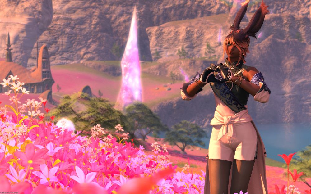 Photo of a Rava Viera character from Final Fantasy XIV standing in a pink flower field with a house and a lake in the background along with a big crystal. The character is making a love heart pose with their hands.