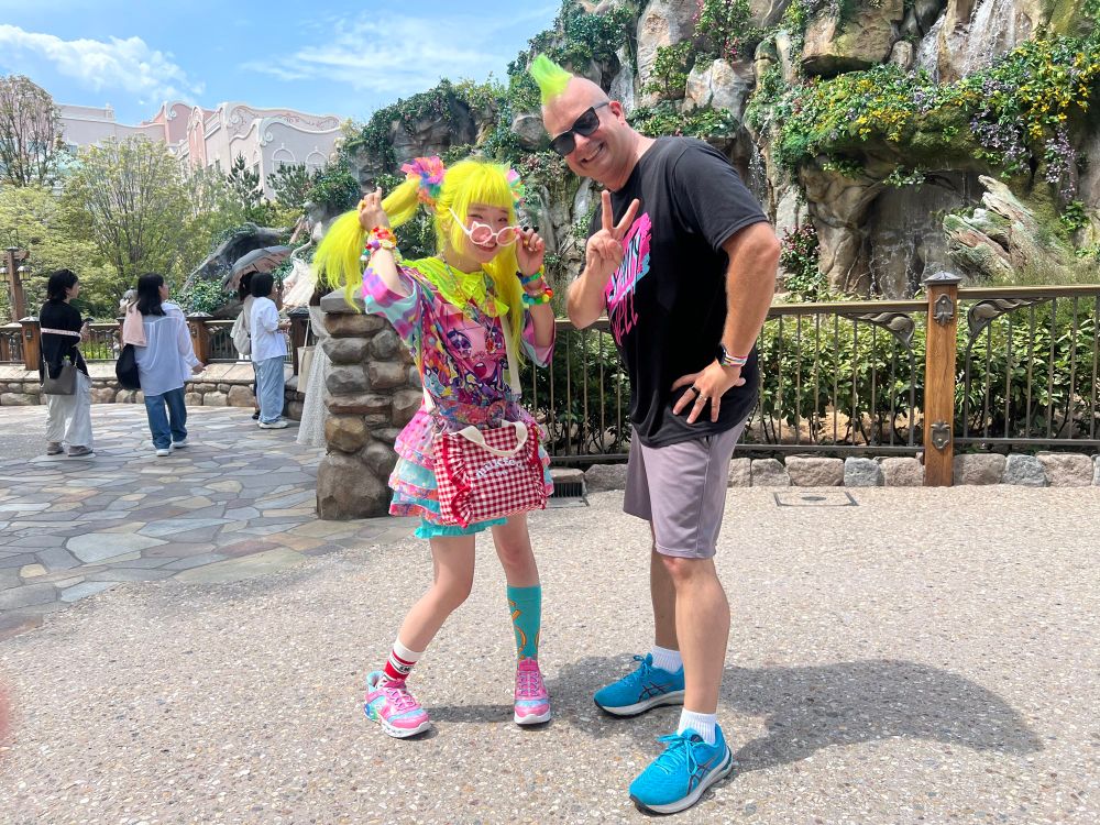 A wild looking Japanese woman dressed in a chaotic Harajuku-style outfit with bright neon yellow hair next to a boring American with a bright neon green Mohawk. 
