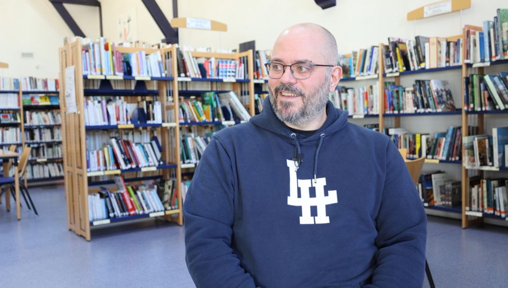 François Dubuc, un homme à lunettes et à barbe avec un hoodie, dans une bibliothèque. Il est assis et sourit en regardant à sa droite, vers le hors champ