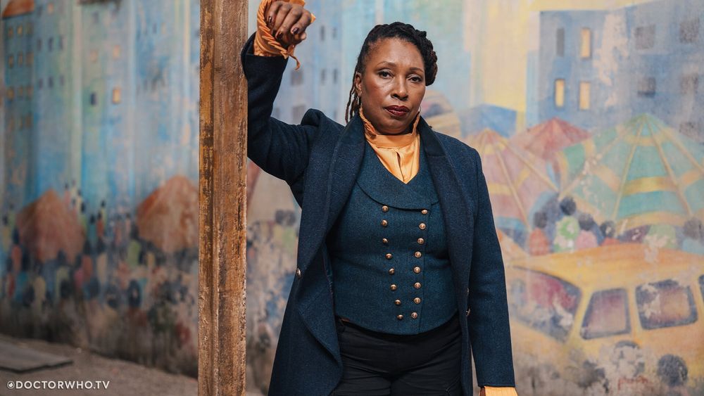 Le Fugitive Docteur, jouée par Jo Martin, une femme noire aux cheveux tressés, avec une longue veste et un gilet aux multiples boutons sombres ainsi qu'une chemise soyeuse orange clair