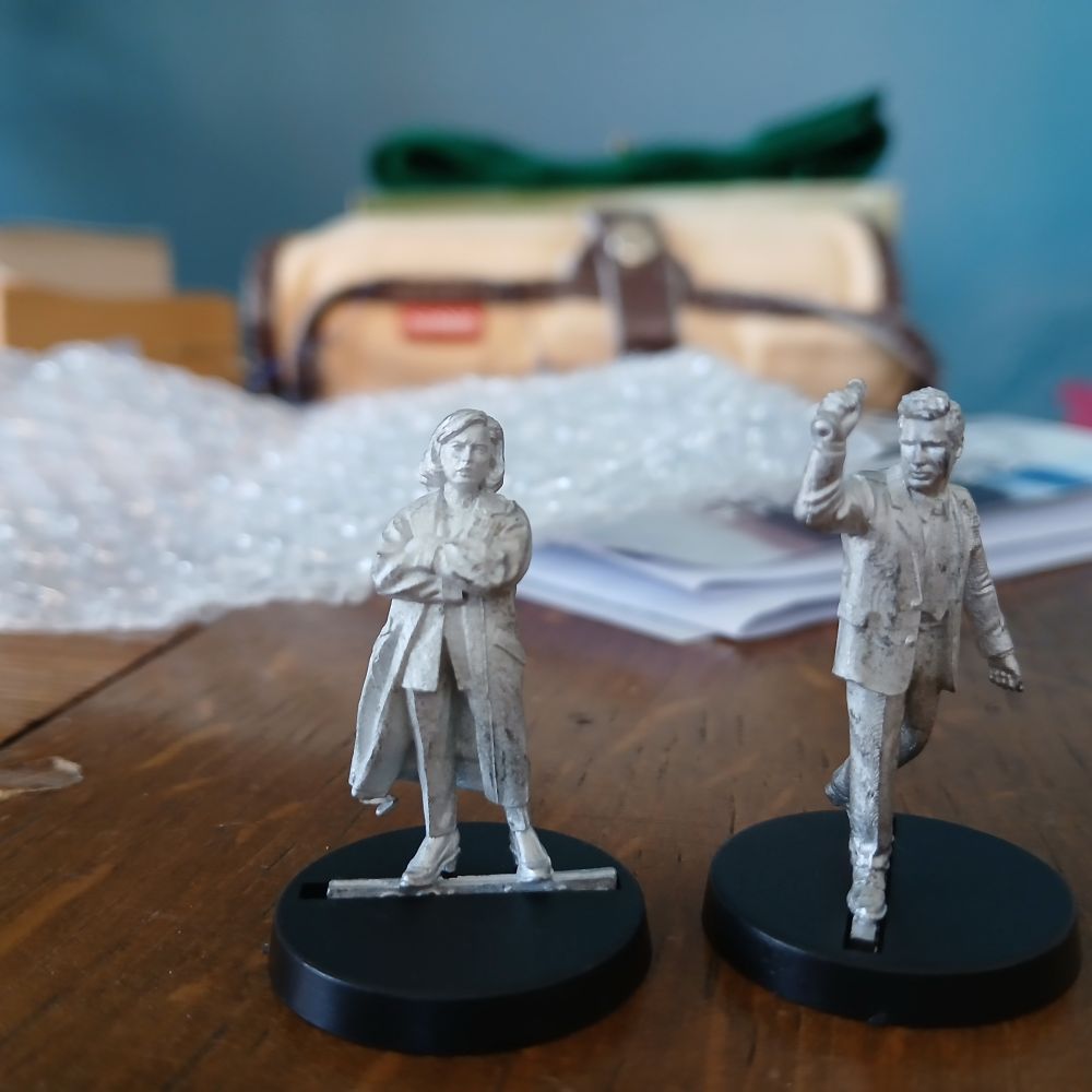 Two unpainted white metal 28mm figurines, FBI Agents from Crooked Dice Games, bearing an uncanny resemblance to Dana Scully and Fox Mulder, but of course legally distinct.