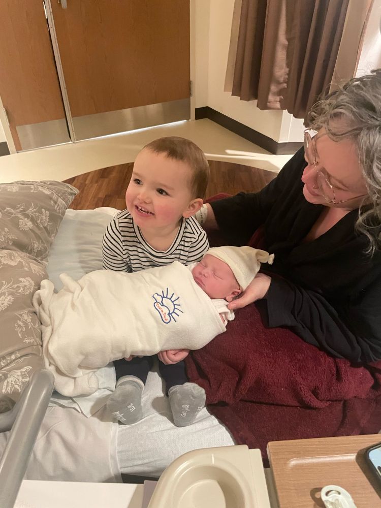 Dolan Emory (2 yo), holds baby brother Oskar Emory (12hrs old) while smiling in delight. Mother Erin Emory lovingly attends to them both. On a hospital bed in a labor & delivery recovery room.