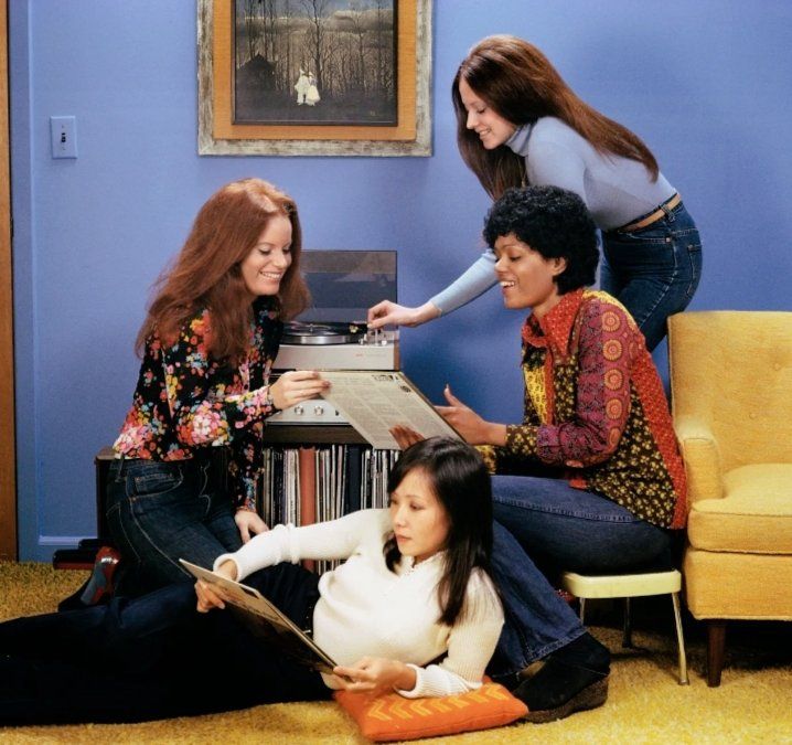 Stock photo of four women listening to records. The room has a lavender wall, and the seating is yellow, with yellow carpet.