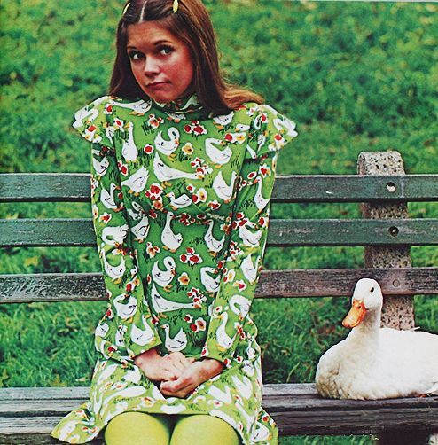 1970s Image of a woman sitting next to a white duck on a park bench. She's wearing a green dress with a white duck print and green tights.