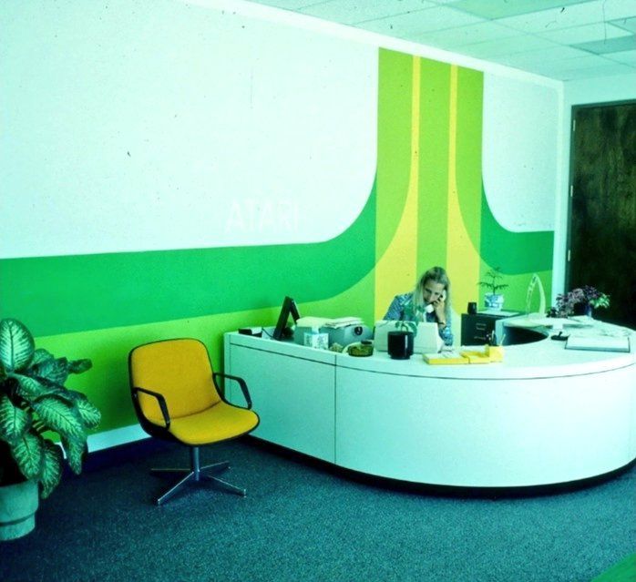 Reception area with a curved white front desk, yellow chair and plant. The wall has a huge yellow and green supergraphic of the Atari Fuji logo.