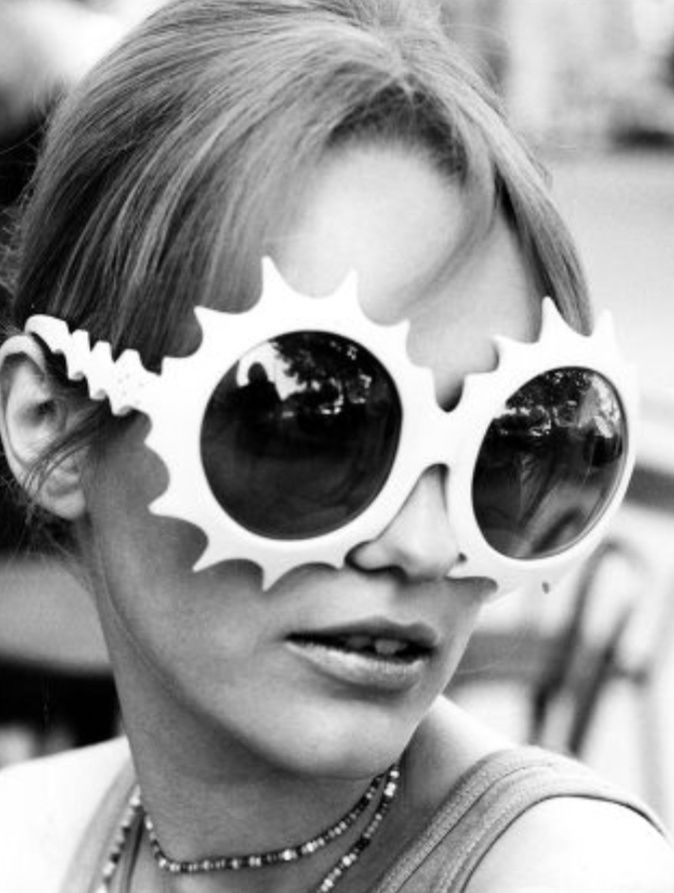 In this photo by Larsen, a woman is wearing white sunglasses shaped like sunbursts by an unknown designer. Novelty eyewear was popularized during this time by the designs of Oliver Goldsmith.