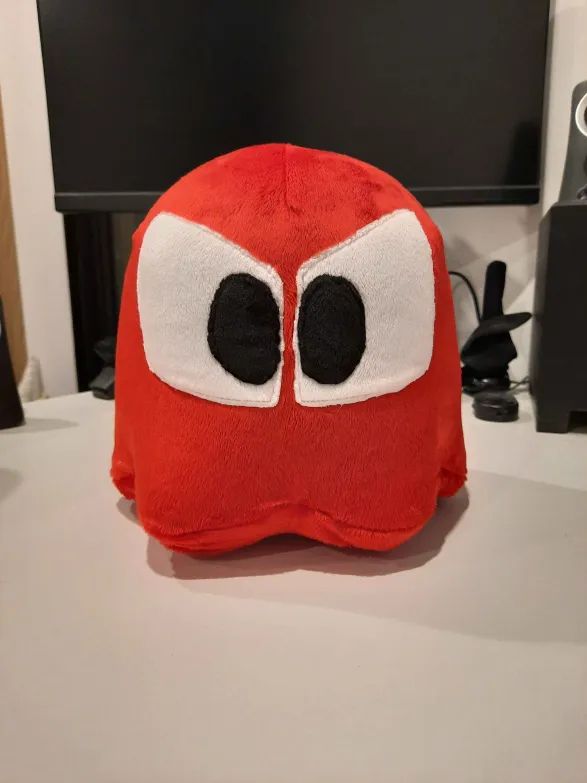A homemade plushie of the red Ghost from PAC-MAN, sitting on my desk facing forward with the camera level to him.