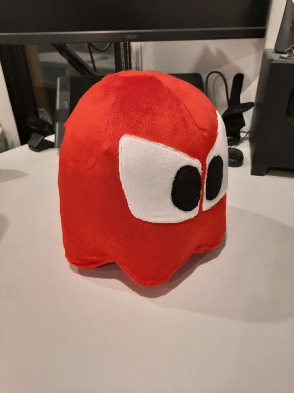A homemade plushie of the red Ghost from PAC-MAN, sitting on my desk facing off to the right with the camera above and angled down.