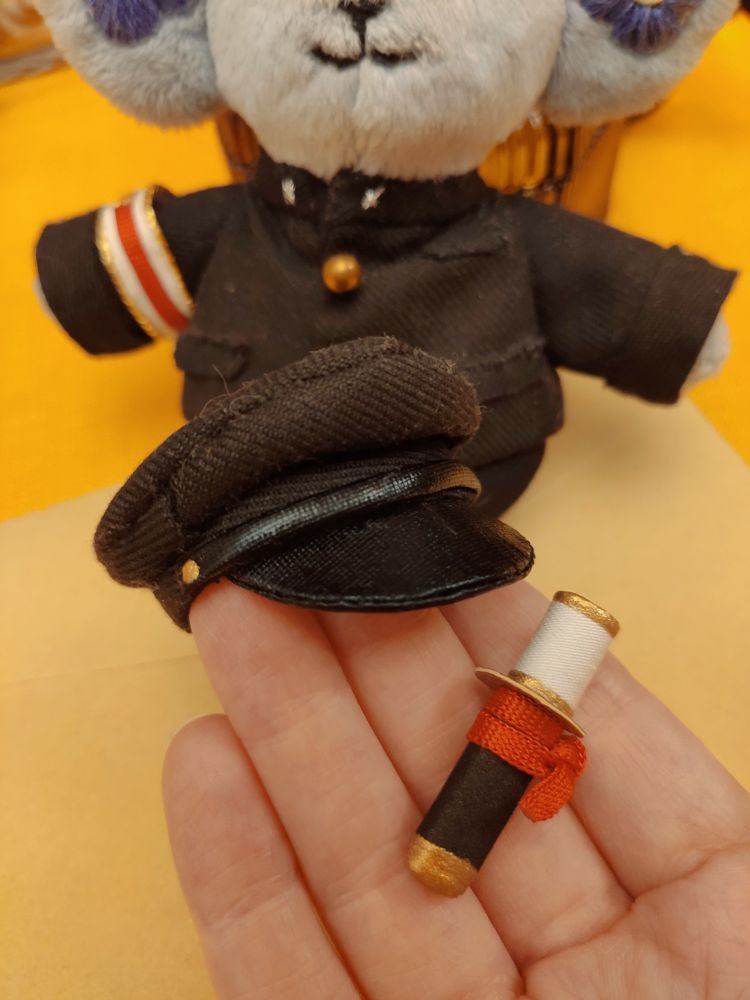 Photo of my hand holding a tiny Japanese student black hat and a tiny crafted katana. The katana has a red ribbon tied and golden details. 