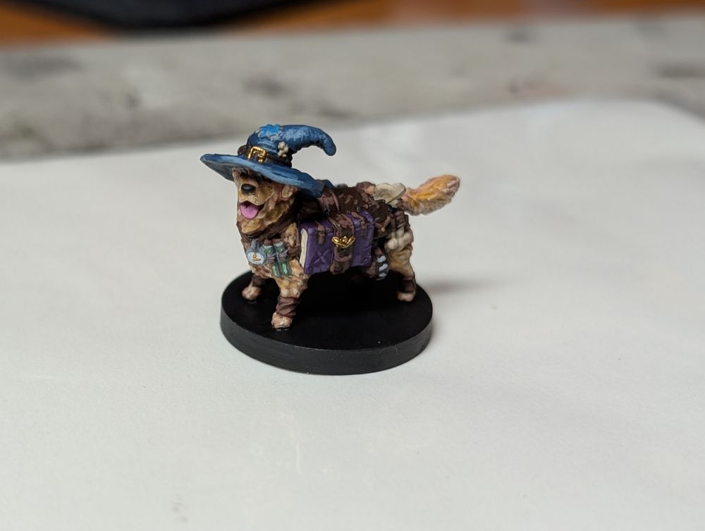 A small painted miniature of a golden retriever wearing a blue wizard hat. He is also wearing a harness that has two books, a lamp, a pair of test tubes, a vial of something green and a watch