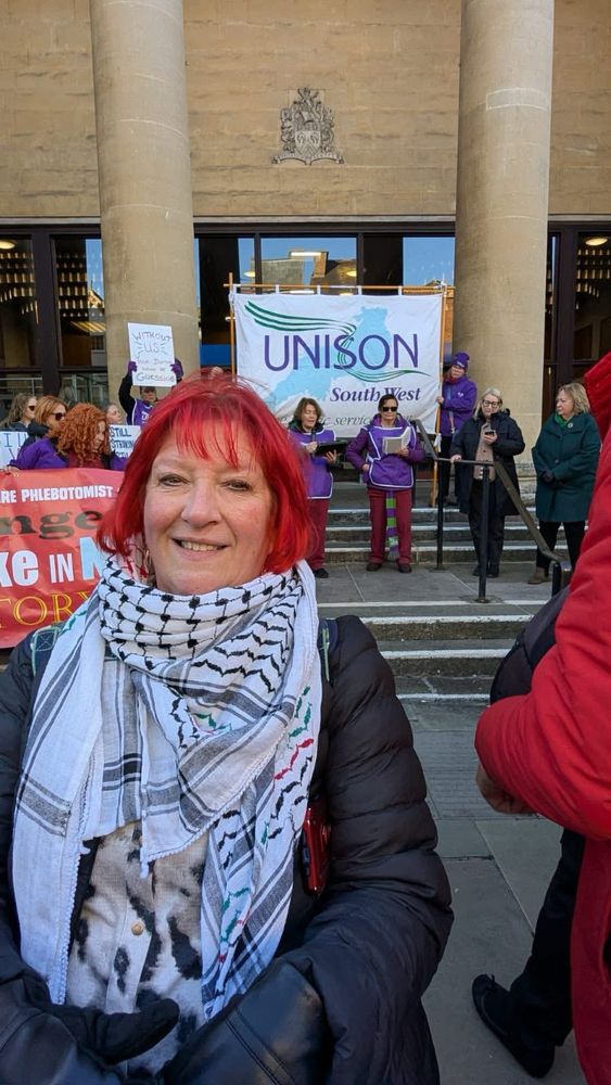 Photo of Andrea Egan (white woman with red hair, dressed in a black coat with keffiyeh) standing alongside striking UNISON workers. Andrea took annual leave to support picket lines in the longstanding industrial action by phlebotomists in Gloucester seeking a regrade from band 2 to band 3. 