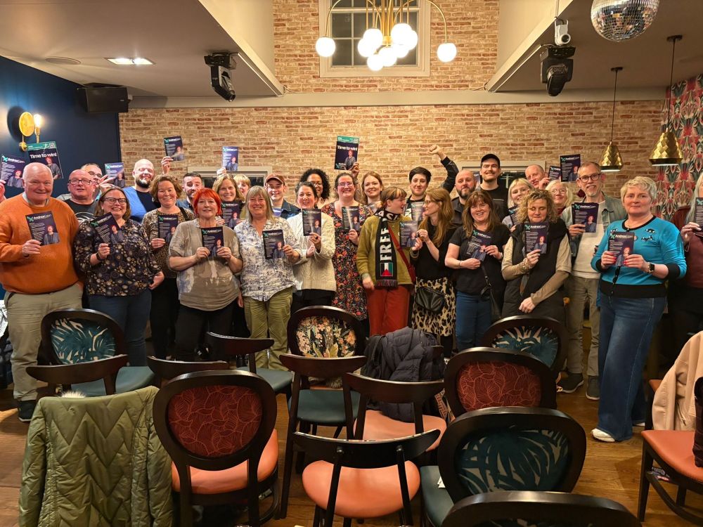 Photo of a room full of smiling people holding up leaflets campaigning for Andrea Egan for General Secretary. Andrea with her distinctive red hair is in the front row. 