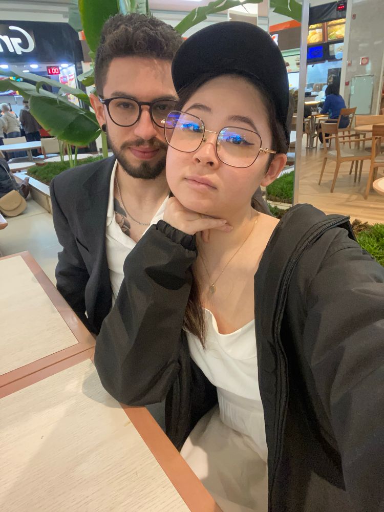 Selfie minha e do Lucas sentados em uma mesa. Estamos olhando pra câmera sérios. Ele usa camisa branca abotoada e blazer preto. Eu uso um vestido branco. Blusa preta
