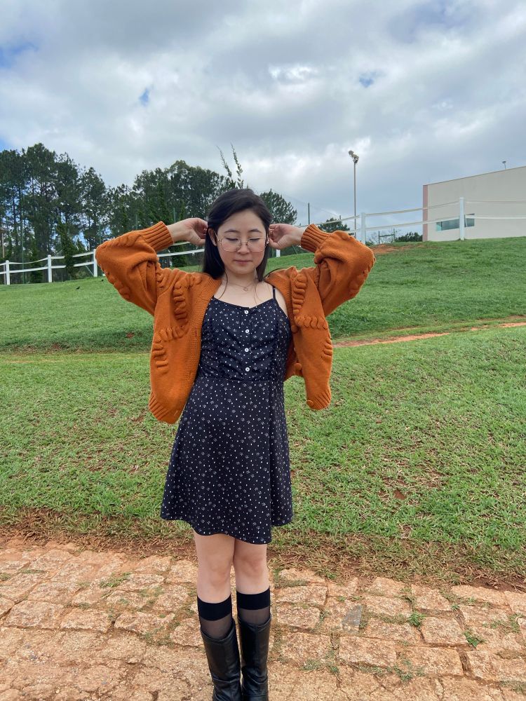 Foto minha em pé levantando os braços mexendo no cabelo e olhando pra baixo. Uso o vestido preto de flores brancas e a blusa alaranjada com bota preta