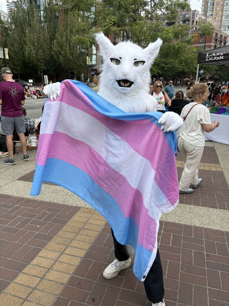 A photo of a person in a white, semi-realistic, canine-like, partial fursuit holding up a trans flag. They are looking at the camera with their mouth open. They stand on a busy sidewalk.
