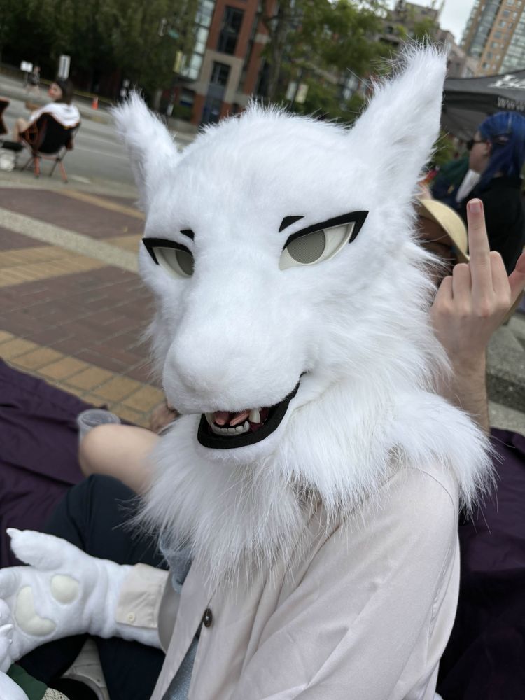 A photo of the same person, looking at the camera while sitting with their mouth open happily. Behind them a friend holds up a middle finger.