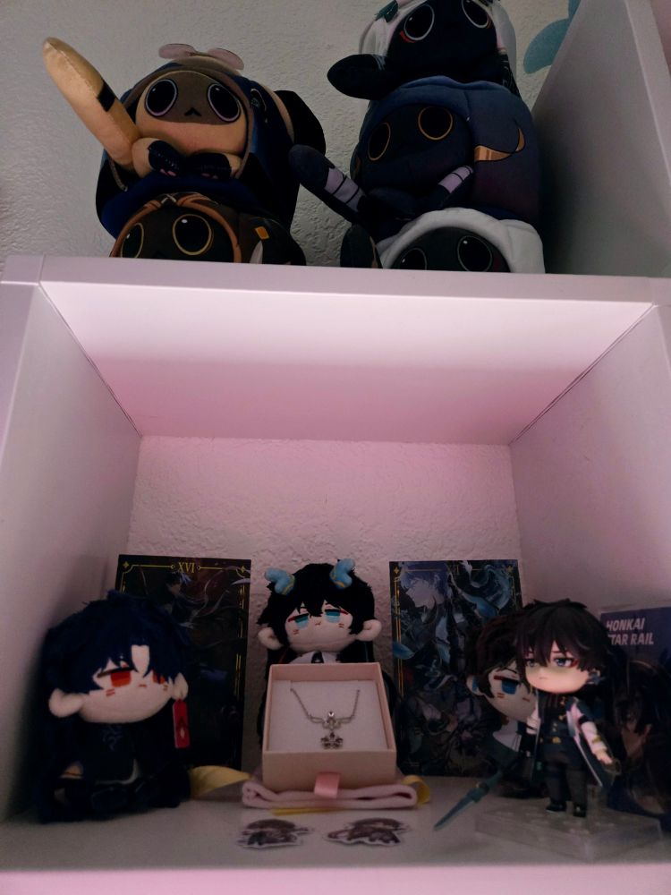 A small shelf with plushies of blade, dan heng, and dan heng in his dragon form. Also has a small chibi figure of Dan Heng, a red necklace, stickers of blade and dan heng from avidyae, and cat cakes that look like the mentioned characters stacked on top of the shelf.