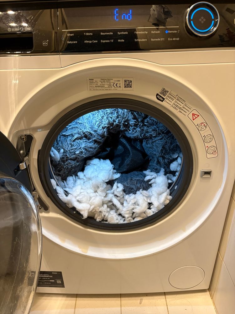 Eine offene Waschmaschine in der ein graues wuscheliges hundebett ist, aber auch ganz viel weißes Füllmaterial in der Trommel liegt 