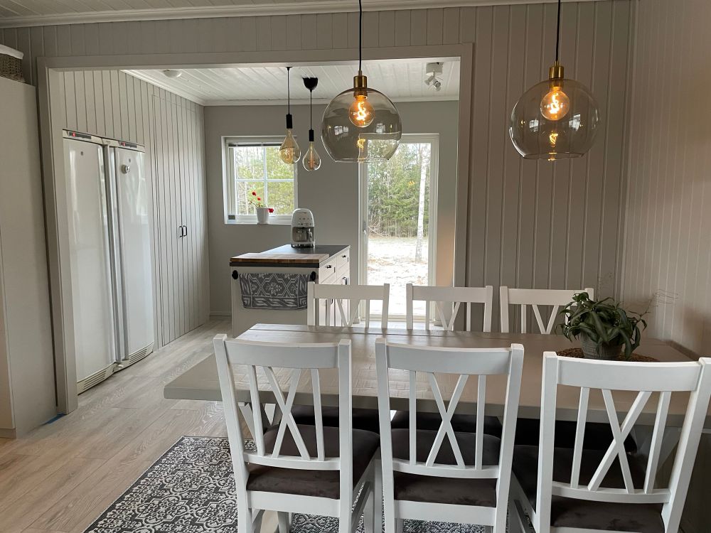 Ytterligare en bild på det nymålade köket, nu med fokus på matplatsen i förgrunden som består av ett rektangulärt bord och sex stycken vita stolar som har en brunaktig sammetssits. I bakgrunden ses delar av köksdelen med kyl, frys och skafferi bakom dubbeldörr i beige med svarta avlånga handtag. Kyl och frys är inbyggd i samma panel som skafferiet. 