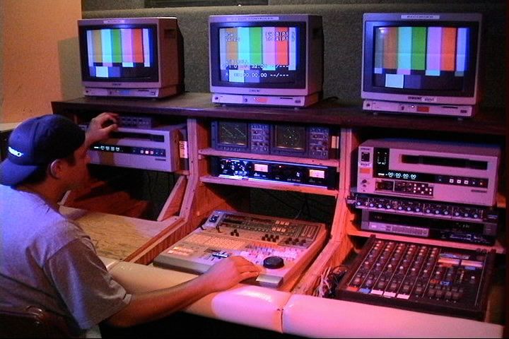 A man sitting at an editing booth with two tape decks, an edit controller, three monitors showing test bars, and an analog sound mixer.
