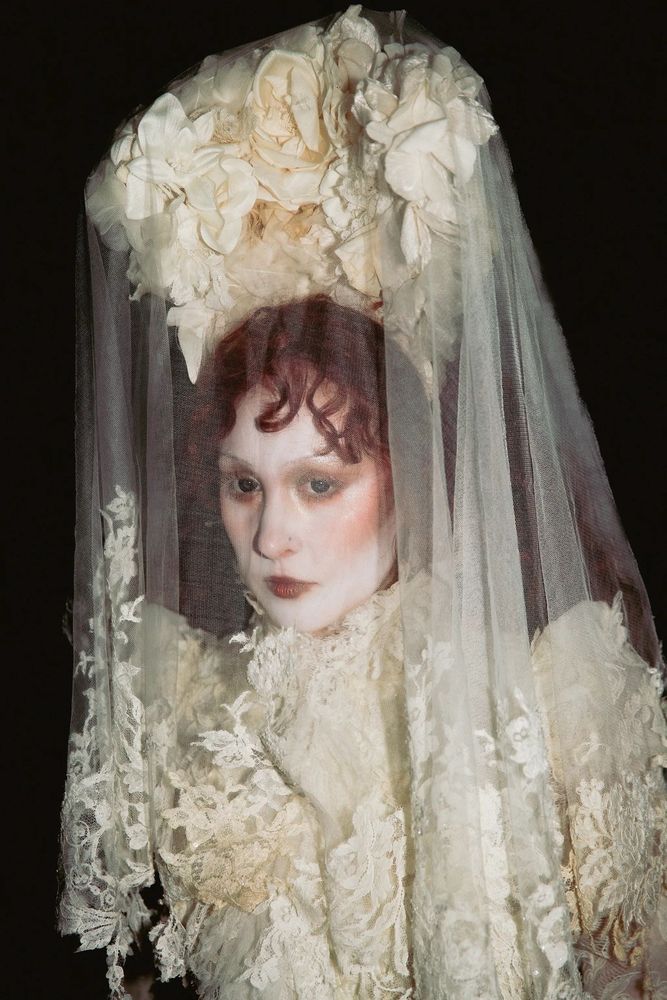 chappell roan backstage at corona capital 2025 in mexico city, MX. photographed by raganhenderson on ig. november 15th, 2025

She is wearing a floral white gown with a long matching veil. Her makeup is a pale white base, dark eyeshadow, bold bronze blush, thin eyebrows and dark lipstick centered in the middle of her lips. She has black eye contacts in and her hair is long, voluminous and curly 