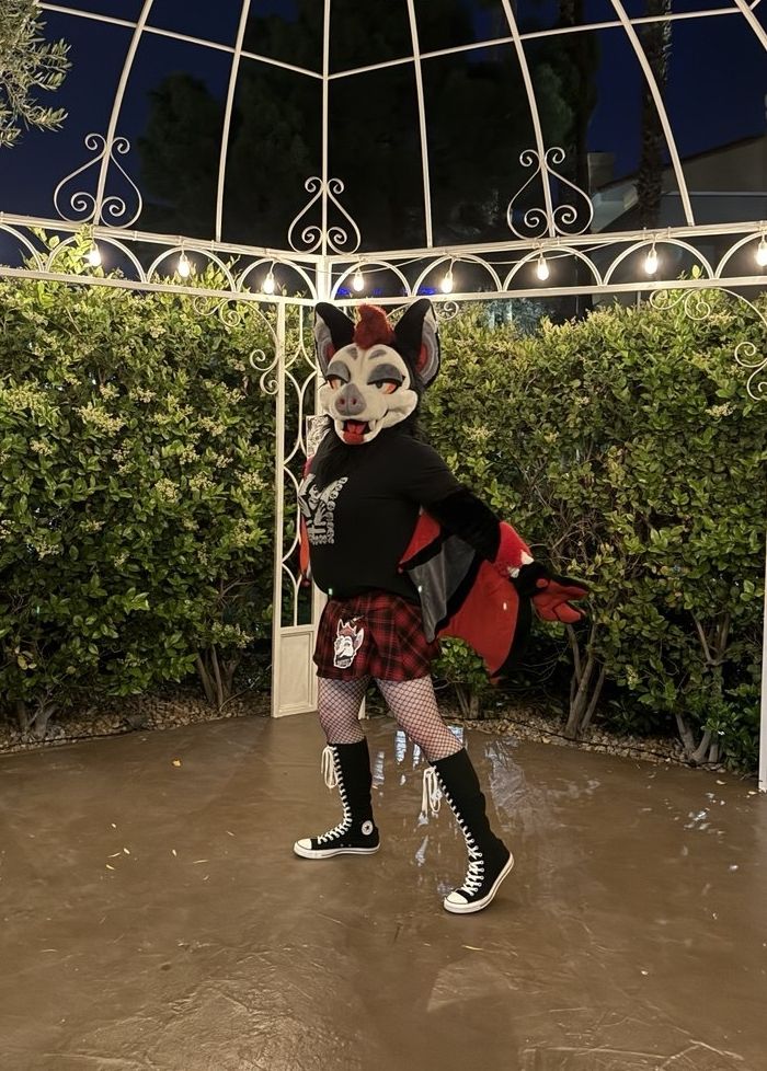Bat partial fursuiter posed in an outdoor gazebo at LVFC2025. She is wearing a black tee-shirt, plaid skirt, fishnets, and knee high Converse boots. 