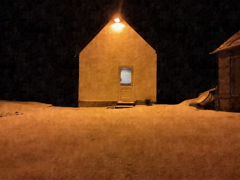The photo shows a small, simple house at night, illuminated by a single warm light above the door. The house has a triangular roof and a plain, light-colored facade. The door, which has a small window, is centered on the wall. The surrounding area is covered in a thin layer of snow, reflecting the warm glow of the light. The background is almost completely dark, making the house stand out starkly. To the right, there is part of another building visible, along with what appears to be a small boat or sled leaning against it. The scene has a quiet, isolated, and slightly mysterious atmosphere, typical of the remote and rugged beauty of the Isle of Skye.