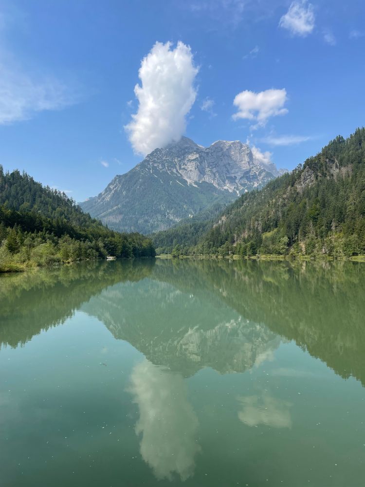 View across a reservoir to Mount Buchstein (and its reflection on the water) on a very sunny day with a white cloud above the summit.