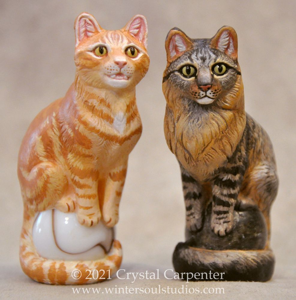 Two cat figurines. One is painted as an orange tabby sitting on a white marble, the other is a longhair brown tabby on a stone marble.