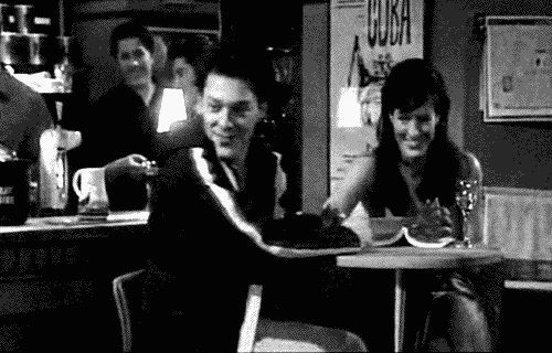 A man, Jeff, and woman are at a table in a bar. The man gives two thumbs up and says "breasts" to another man at a separate table, who does the same in response. 