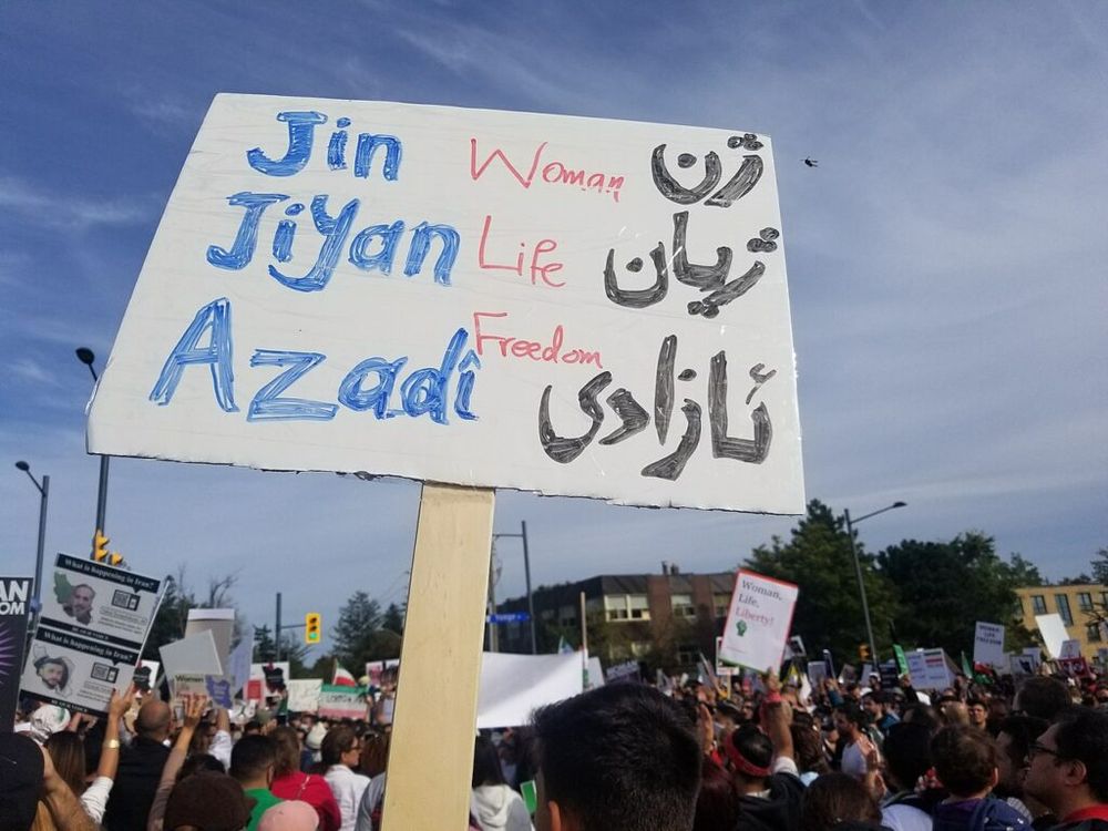 Picture of a protest in Iran with a cardboard saying the slogan "Jin Jiyan Azadî" in multiple languages/scripts