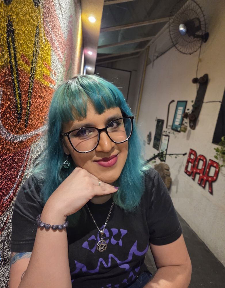 Foto que a Aurora tirou de mim. Sou uma mulher branca de cabelos verdes na altura do ombro, camiseta preta e a mão está abaixo do queixo.