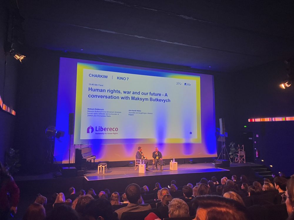 Ein gut besuchtes Panelgespräch in einem dunklen Kinosaal mit zwei Personen auf einer beleuchteten Bühne. Im Hintergrund ist eine große Projektionsfläche mit einer Präsentation zu sehen. Der Titel des Panels lautet: 'Human rights, war and our future - A conversation with Maksym Butkevych'. Die Namen der Teilnehmenden, Maksym Butkevych und Jan-Henrik Wiebe, sowie das Logo der Organisation 'Libereco - Partnership for Human Rights' sind ebenfalls sichtbar.