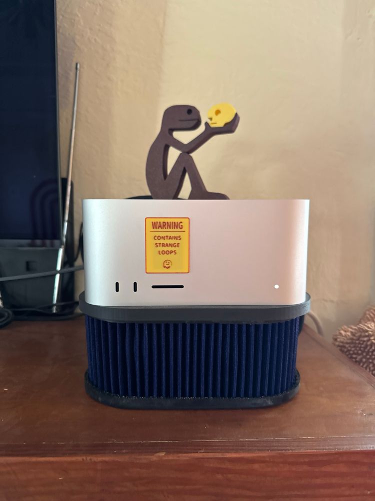 A recursive idol atop a cybernetic altar. A stick-figure Hamlet contemplates a neon skull on the crown of a Mac Studio, which itself rests on a ribbed obsidian-blue plinth like an alien air filter. A warning sigil proclaims: “CONTAINS STRANGE LOOPS,” as if to ward off linear thinkers. The machine hums with the quiet dread of self-awareness. Somewhere, Hofstadter weeps.