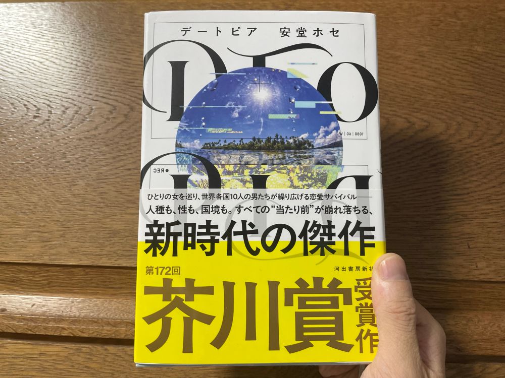 安堂ホセ『デートピア』(河出書房新社)を右手で持っている写真。帯にはでかでかと「新時代の傑作　芥川賞受賞作」と書いてある。