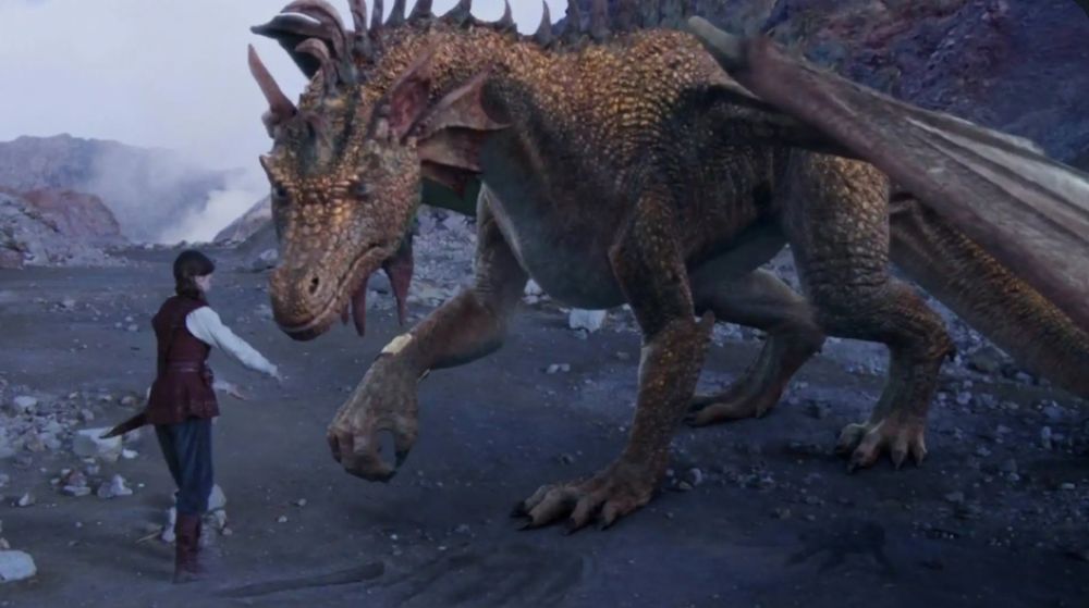 A scene from "Voyage of the Dawn Treader": A large, bronze-scaled dragon with leathery wings stands facing a young man in an open, rocky landscape. The man, dressed in a white shirt and a dark vest, has his hand extended toward the dragon's snout as if in greeting or offering comfort.
